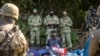 Польскія (на пярэднім пляне) і беларускія (на заднім пляне) памежнікі стаяць побач з мігрантамі, якія, як мяркуецца, паходзяць з Афганістана і сядзяць на зямлі ў невялікай вёсцы Уснаж-Гурны каля Беластока, на паўночным усходзе Польшчы, разьмешчанай недалёка ад мяжы. 2021