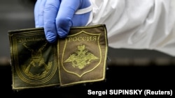 Український криміналіст показує нашивки, знайдені на тілі російського солдата, ексгумованого в селі Завалівка на захід від Києва, коли спецпідрозділ оглядає рефрижераторний вагон із загиблими росіянами, 11 травня 2022 року